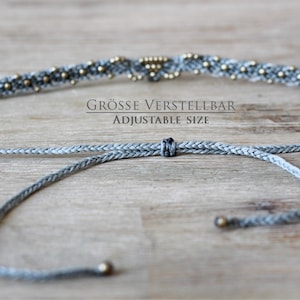 May include: A gray braided cord necklace with gold beads and a sliding knot for adjustable size. The text "GR&Ouml;SSE VERSTELLBAR ADJUSTABLE SIZE" is printed on a wooden surface.