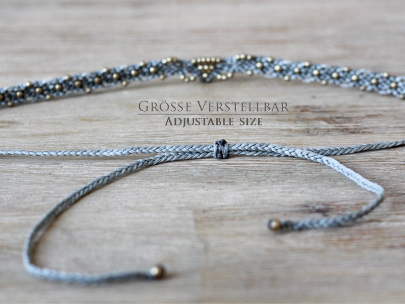 May include: A gray braided cord necklace with gold beads and a sliding knot for adjustable size. The text "GR&Ouml;SSE VERSTELLBAR ADJUSTABLE SIZE" is printed on a wooden surface.