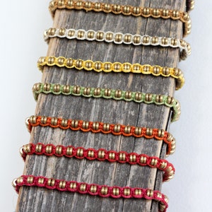 Könnte beinhalten: Ein Set aus zehn Armbändern mit verschiedenen farbigen Schnüren und goldenen Perlen. Die Armbänder sind in einer Reihe auf einer Holzoberfläche angeordnet.
