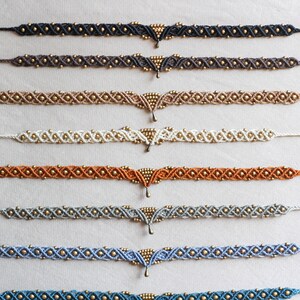 May include: A display of eight handmade macrame anklets with gold beads and a gold pendant. The anklets are in various colors, including black, brown, beige, white, orange, gray, blue, and teal.