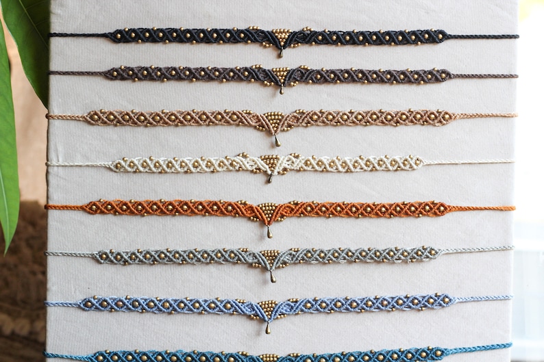 May include: A display of eight handmade macrame anklets with gold beads and a gold pendant. The anklets are in various colors, including black, brown, beige, white, orange, gray, blue, and teal.