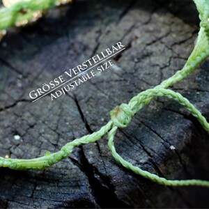 May include: A green braided cord bracelet with gold beads. The bracelet is adjustable and has a knot at the end. The text "GR&Ouml;SSE VERSTELLBAR ADJUSTABLE SIZE" is printed on the bracelet.