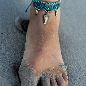 May include: A turquoise and gold ankle bracelet with leaf charms. The bracelet is on a person's ankle, which is covered in sand.