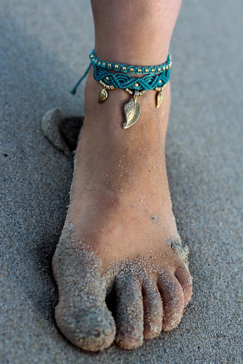 May include: A turquoise and gold ankle bracelet with leaf charms. The bracelet is on a person's ankle, which is covered in sand.