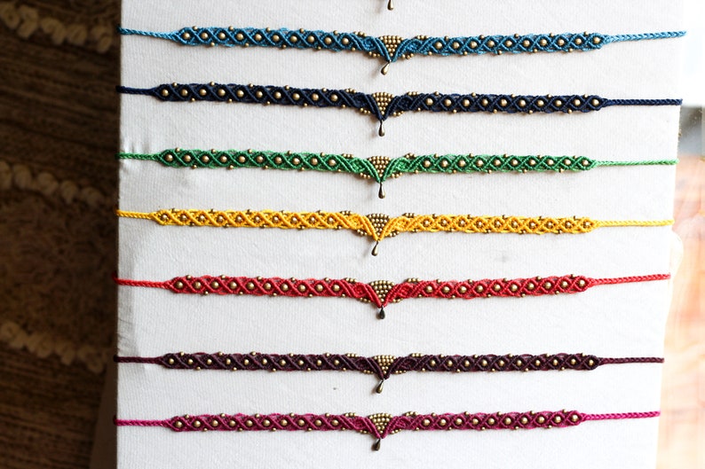 May include: A set of eight colorful friendship bracelets with gold beads and a gold pendant. The bracelets are displayed on a white background.
