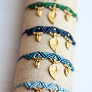May include: A stack of colorful braided bracelets with gold leaf charms. The bracelets are displayed on a beige surface.