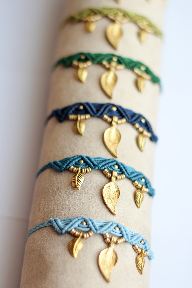 May include: A stack of colorful braided bracelets with gold leaf charms. The bracelets are displayed on a beige surface.
