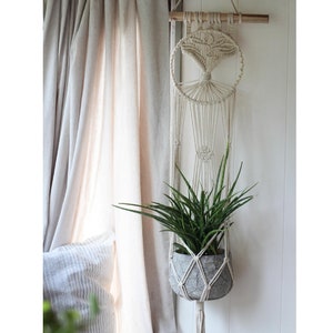 Peut inclure: Un porte-plante en macramé avec un motif circulaire est suspendu à un mur blanc. Une plante verte est dans un pot gris à l'intérieur du porte-plante.