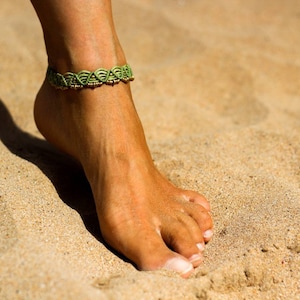 Può includere: Primo piano di un piede femminile nudo con un braccialetto alla caviglia verde e oro su una spiaggia sabbiosa.