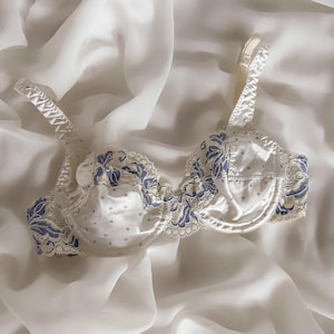 Puede incluir: Sujetador de satén blanco con bordado floral azul y ribete de encaje. El sujetador presenta tirantes ajustables y un diseño delicado. Las copas están adornadas con pequeños patrones florales azules, creando una estética vintage.