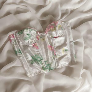 May include: A white satin corset with a floral print featuring pink and green flowers. The corset has boning and a sweetheart neckline.