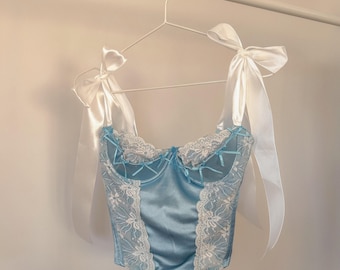 May include: A light blue satin corset with white lace trim and white satin ribbon straps. The corset has a sweetheart neckline and a boned bodice.