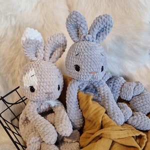 Könnte beinhalten: Zwei gehäkelte graue Plüschhasen mit weißen Akzenten. Die Hasen sitzen auf einer gelben Decke in einem schwarzen Drahtkorb.