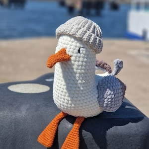 Puede incluir: Una gaviota de crochet blanco con un sombrero de crochet gris y pies de crochet naranja. La gaviota está sentada en una superficie gris.