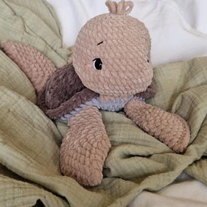 May include: A soft, brown and gray crocheted turtle plush toy with a sweet face. The toy is lying on a green blanket.