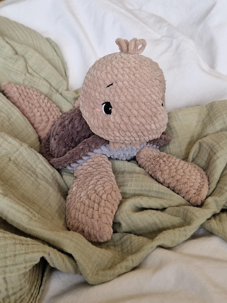 May include: A soft, brown and gray crocheted turtle plush toy with a sweet face. The toy is lying on a green blanket.
