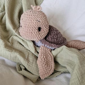 May include: A crocheted plush turtle toy with a brown shell and pink body. The turtle is lying on a green blanket.
