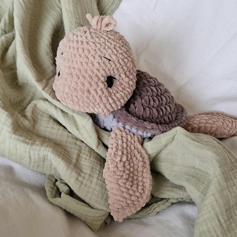 May include: A crocheted plush turtle toy with a brown shell and pink body. The turtle is lying on a green blanket.