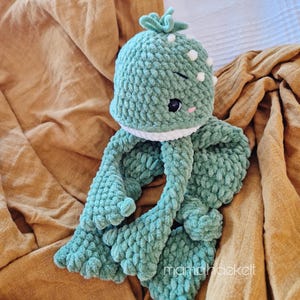 May include: A crocheted green octopus plush toy with white accents and a pink blush on its cheeks. The octopus is sitting on a brown blanket.