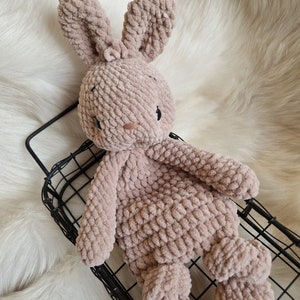 Puede incluir: Un suave juguete de conejo de crochet marr&oacute;n con orejas largas. El conejo est&aacute; sentado en una cesta de alambre negro.