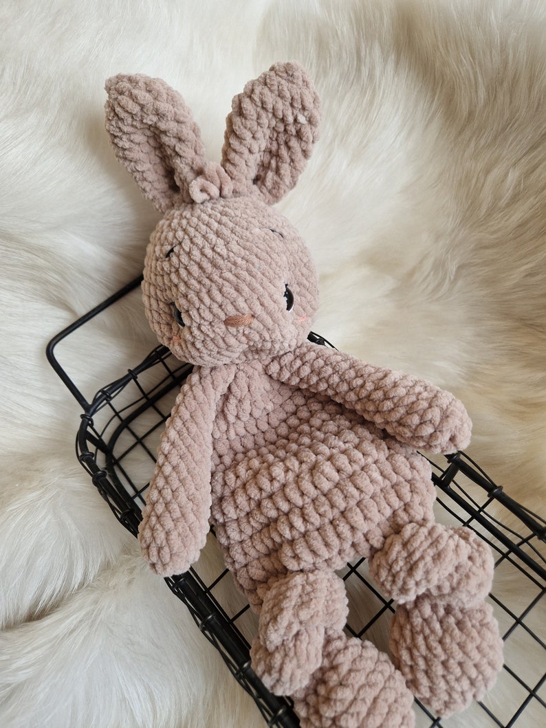 Puede incluir: Un suave juguete de conejo de crochet marr&oacute;n con orejas largas. El conejo est&aacute; sentado en una cesta de alambre negro.