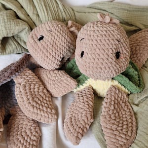 May include: Two crocheted plush toys in the shape of a turtle. The turtle on the left is brown and has a simple design. The turtle on the right is brown with a green shell and a yellow belly.