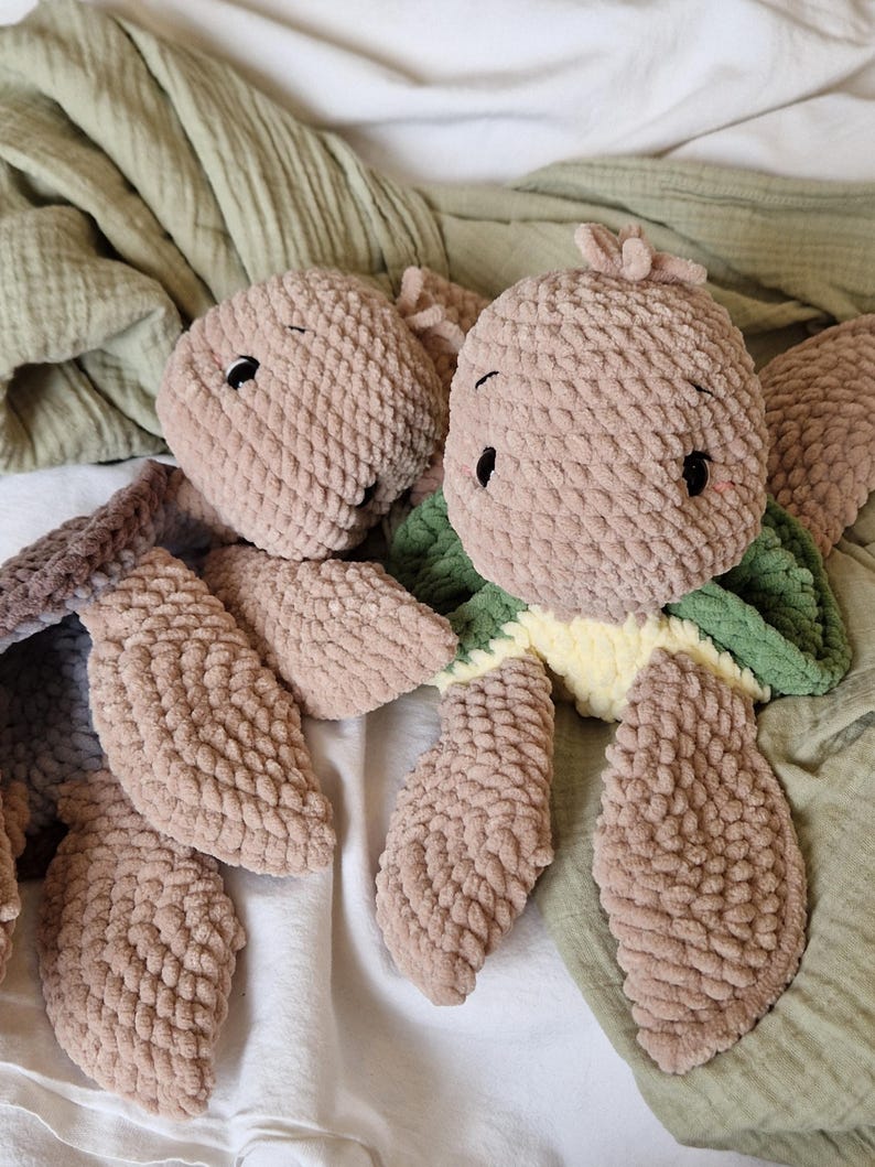 May include: Two crocheted plush toys in the shape of a turtle. The turtle on the left is brown and has a simple design. The turtle on the right is brown with a green shell and a yellow belly.
