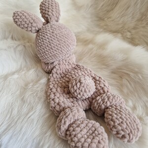 Puede incluir: Un suave juguete de peluche de conejo de crochet en color rosa claro. El conejo tiene orejas largas, una cola peque&ntilde;a y un cuerpo rechoncho.