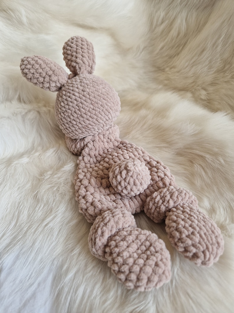 Puede incluir: Un suave juguete de peluche de conejo de crochet en color rosa claro. El conejo tiene orejas largas, una cola peque&ntilde;a y un cuerpo rechoncho.