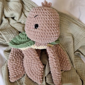 May include: A plush crocheted turtle toy with a green shell and brown body. The toy has a cute face with black eyes and a stitched smile.