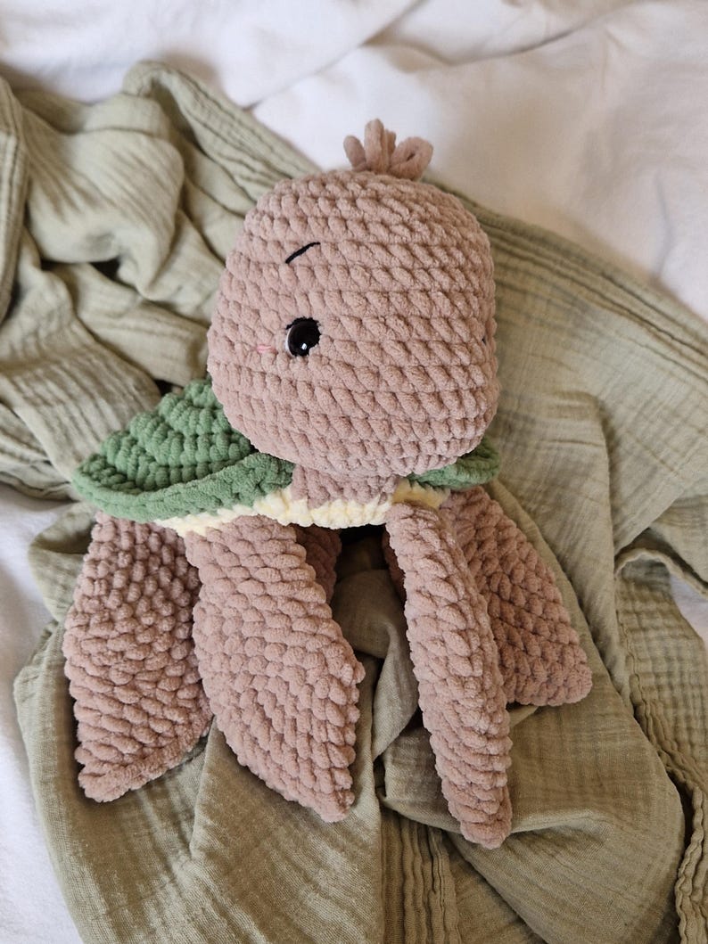 May include: A plush crocheted turtle toy with a green shell and brown body. The toy has a cute face with black eyes and a stitched smile.