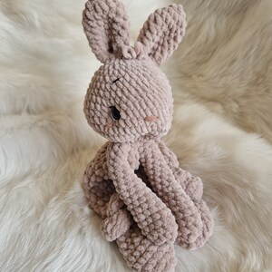 Puede incluir: Un juguete de peluche de conejo de crochet de color marr&oacute;n claro. El conejo est&aacute; sentado sobre una superficie blanca y esponjosa.