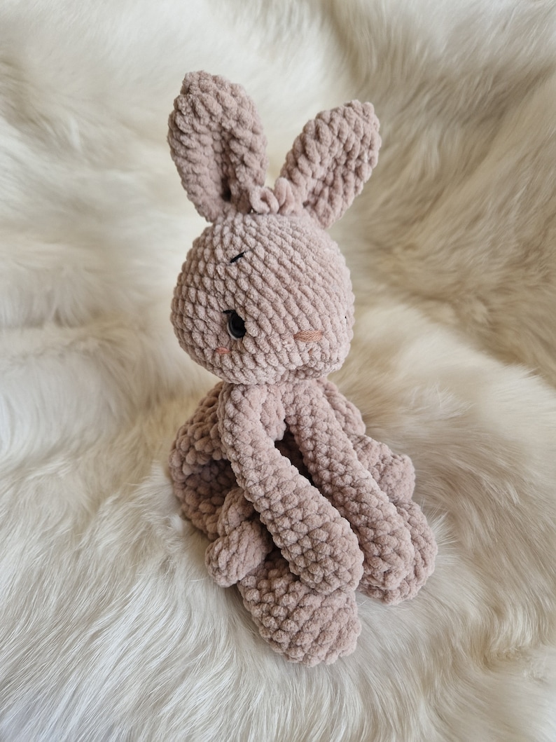 Puede incluir: Un juguete de peluche de conejo de crochet de color marr&oacute;n claro. El conejo est&aacute; sentado sobre una superficie blanca y esponjosa.