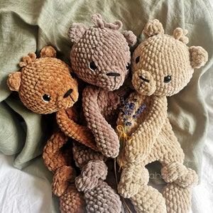 Puede incluir: Tres osos de peluche tejidos a crochet en colores marrón, beige y gris. Los osos están sentados sobre un fondo de tela blanca y verde.