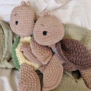 May include: Two crocheted plush turtle toys, one brown and one beige, with green and yellow crocheted shells. The turtles are lying on a green and white blanket.