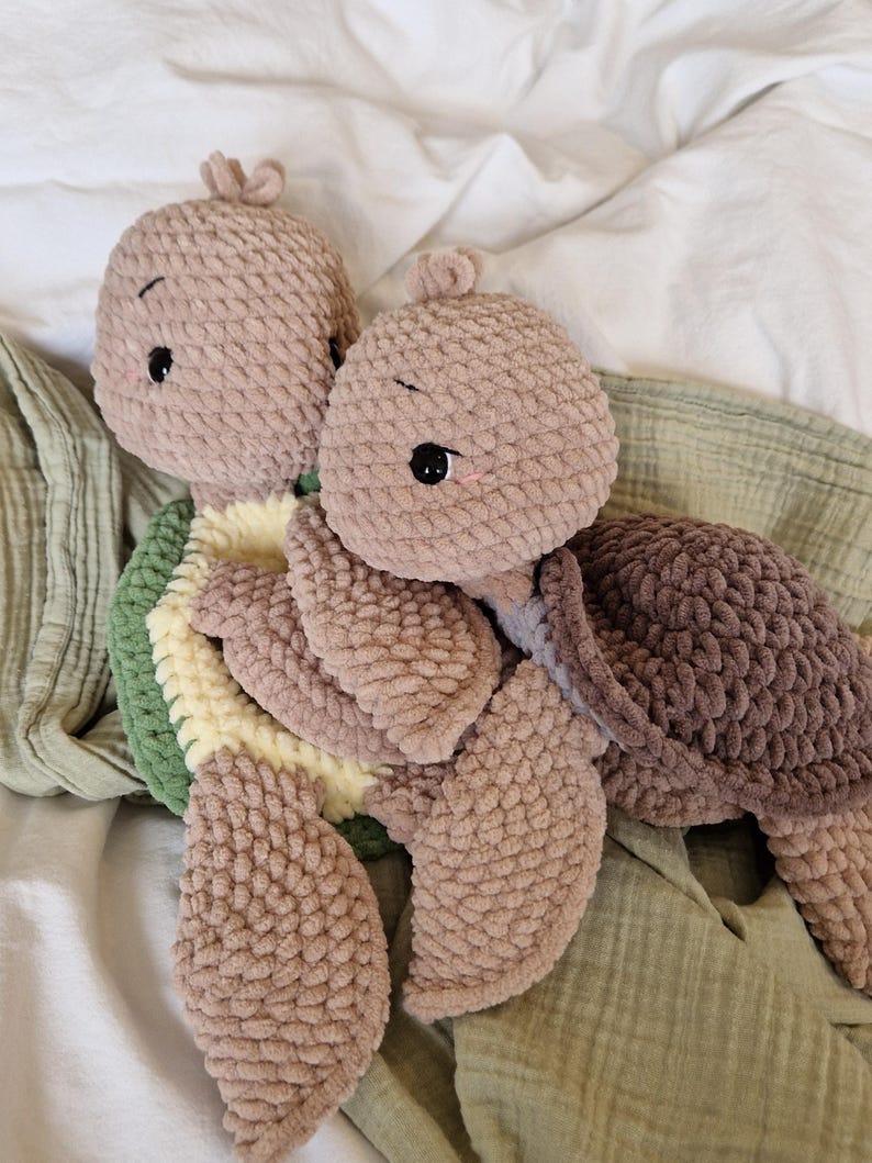 May include: Two crocheted plush turtle toys, one brown and one beige, with green and yellow crocheted shells. The turtles are lying on a green and white blanket.