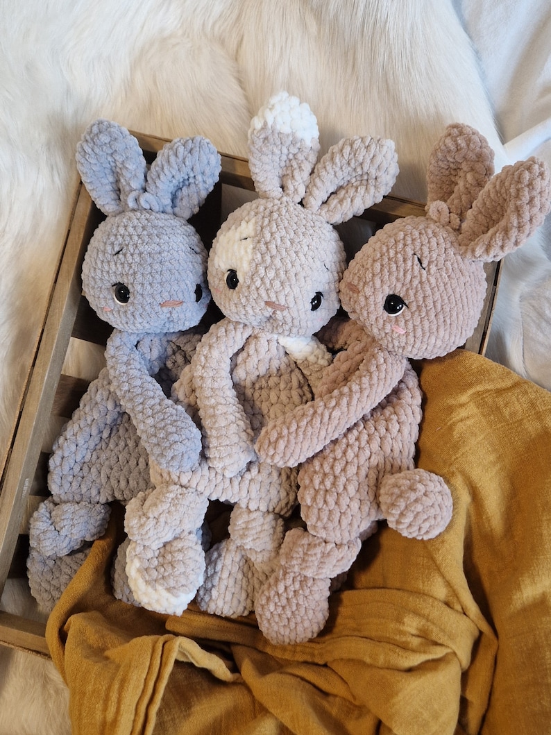 Puede incluir: Tres peluches de conejo de crochet en una caja de madera. Un conejo es gris claro, otro es marr&oacute;n claro y el &uacute;ltimo es una mezcla de marr&oacute;n claro y blanco.