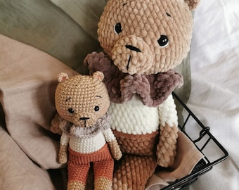 May include: Two handmade crochet teddy bears. The larger bear is light brown with a dark brown collar. The smaller bear wears a scarf, white shirt, and orange trousers. Both bears have black eyes and are displayed in a black wire basket.