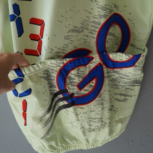 May include: A light green cycling jersey with a blue and red graphic design. The jersey has a pocket on the back and features a digital display with the number "33" on the front.
