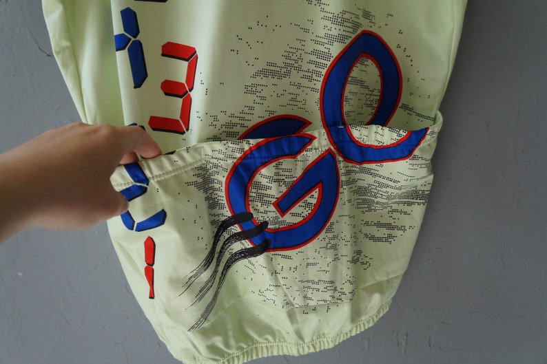 May include: A light green cycling jersey with a blue and red graphic design. The jersey has a pocket on the back and features a digital display with the number "33" on the front.