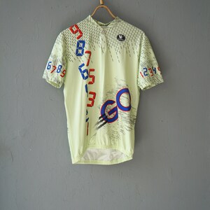 May include: A light green short-sleeved cycling jersey with a white zipper and a blue and red "GO" graphic. The jersey has a pattern of diagonal lines and numbers on the front.