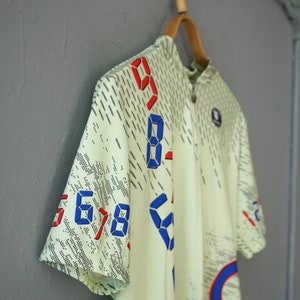 May include: A light green short-sleeved cycling jersey with a zipper closure. The jersey has a digital print design with blue and red numbers.