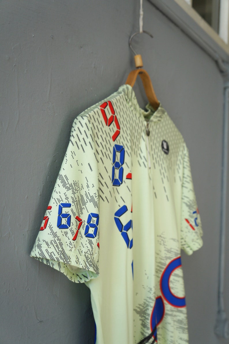 May include: A light green short-sleeved cycling jersey with a zipper closure. The jersey has a digital print design with blue and red numbers.