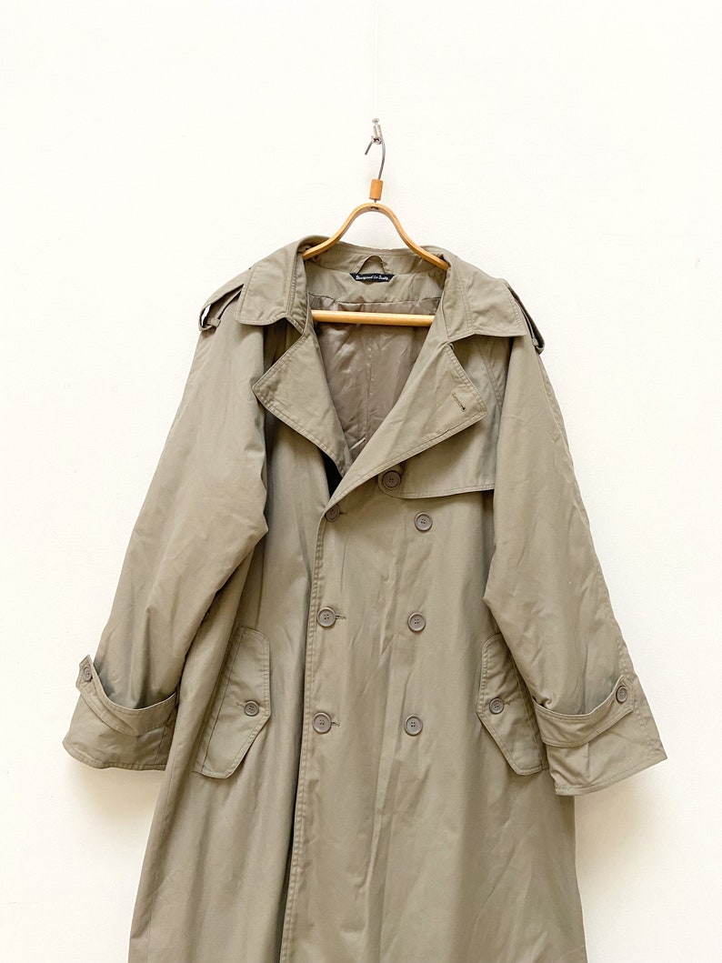 May include: A long, beige trench coat with a double-breasted closure and a belt. The coat has a classic design with a collar, pockets, and a buttoned cuff.