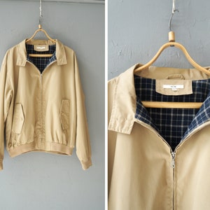 Könnte beinhalten: Beige Reißverschlussjacke mit Kragen und zwei Vordertaschen. Die Jacke hat ein blau-schwarz kariertes Futter und hängt an einem Holzbügel vor einer grauen Wand. Der Stil der Jacke ist klassisch und lässig.