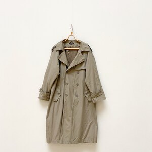 May include: A long, beige trench coat with a double-breasted closure and a belt. The coat has a classic design with a collar, pockets, and a back vent.