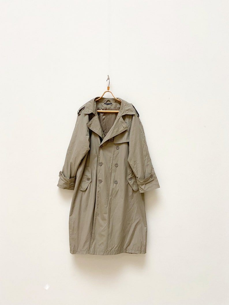 May include: A long, beige trench coat with a double-breasted closure and a belt. The coat has a classic design with a collar, pockets, and a back vent.