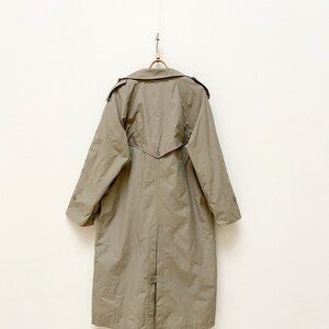 May include: A long, khaki-colored trench coat with a button closure and a belt in the back. The coat is hanging on a wooden hanger against a white wall.
