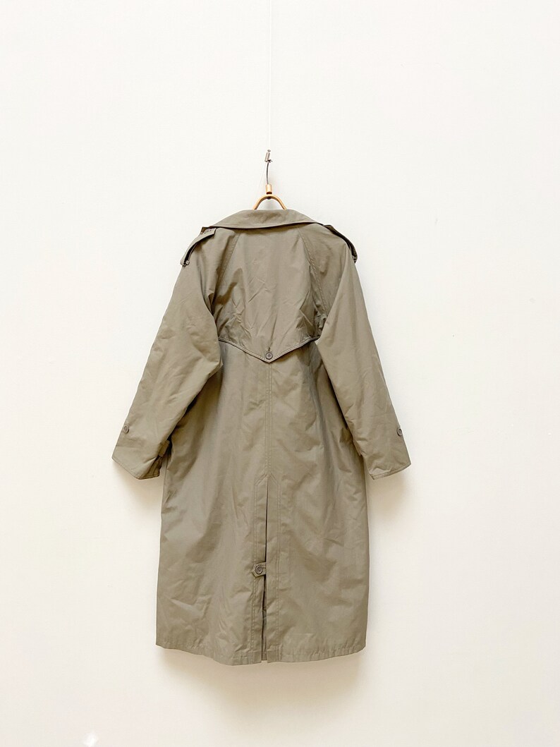 May include: A long, khaki-colored trench coat with a button closure and a belt in the back. The coat is hanging on a wooden hanger against a white wall.