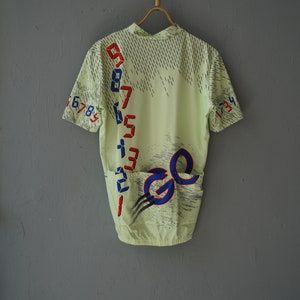 May include: A light green cycling jersey with a blue and red graphic design. The design features a number sequence and the letter "G" repeated twice.